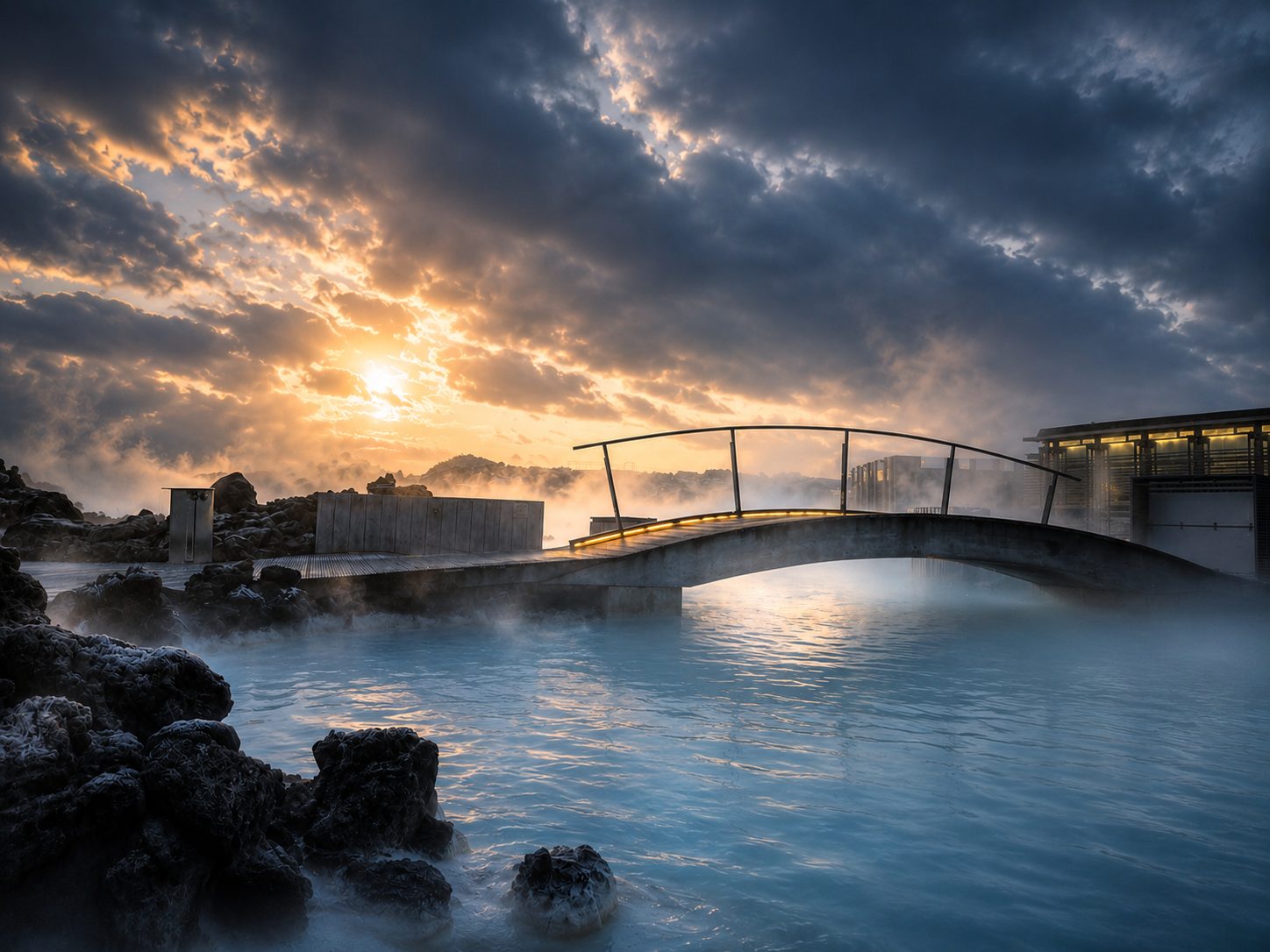 A footbridge over milky blue geothermal water under a dramatic sky